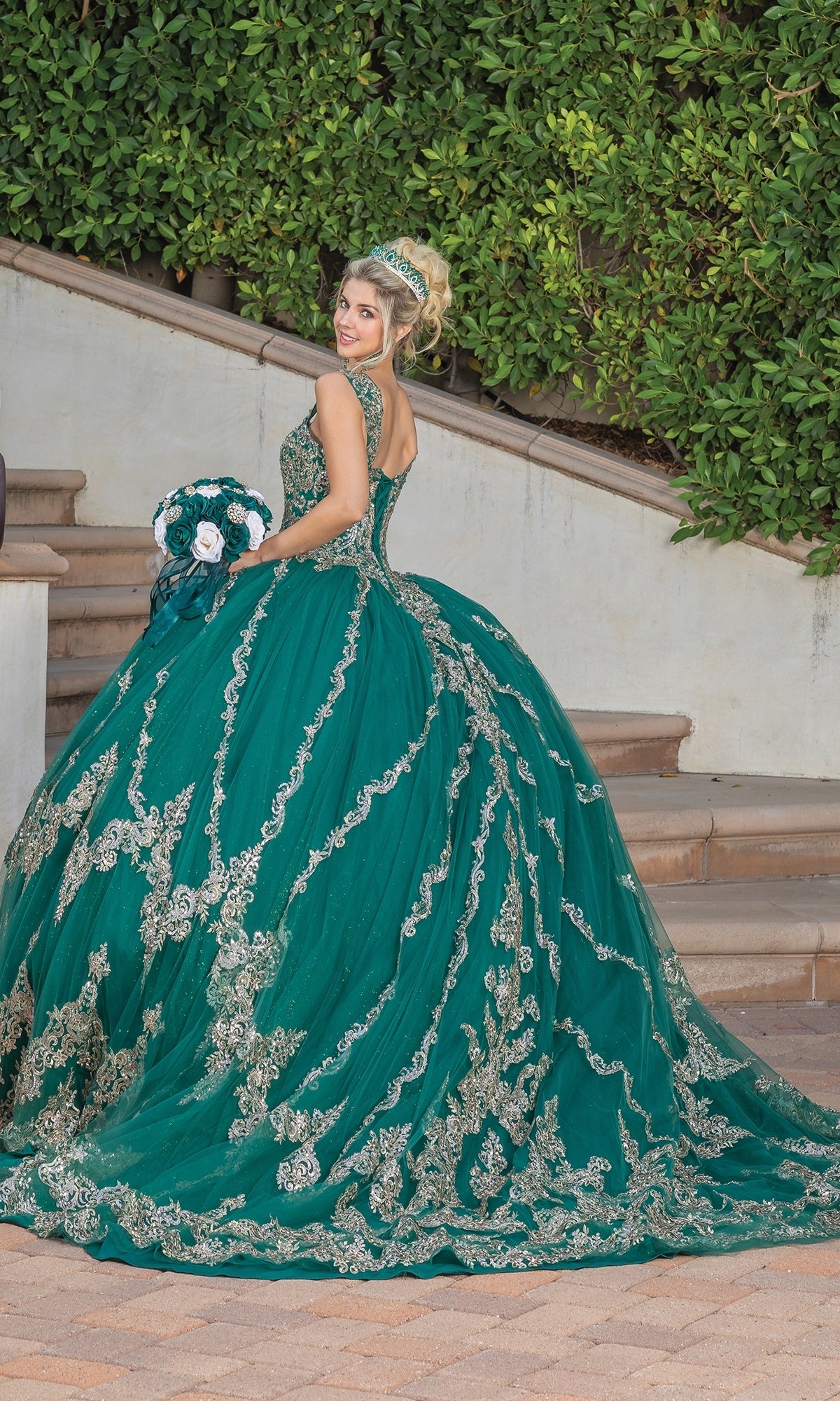 A woman in the Quinceanera Dress 1779 by Dancing Queen, featuring a hunter green ball gown and lace embroidered sweetheart neckline, stands on an outdoor staircase with a matching bouquet and tiara.